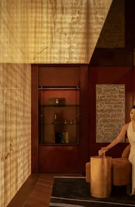 A woman sits on a chair in a cozy living room with a couch, clocks, and a vase, surrounded by warm lighting and subtle shadows