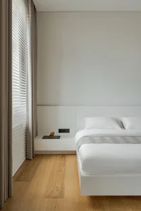 A calm and inviting bedroom with a white bed, hardwood floor, and a few personal items such as books and a remote control