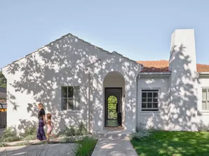 A white house with a large doorway, porch, and small tree, with two women walking towards the entrance