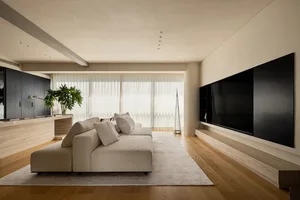 A spacious living room with a large couch, two chairs, a mounted television, and a potted plant, set against a backdrop of white walls and natural light