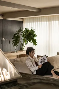 A woman reads a book on a couch in a cozy living area with a potted plant, vases, and a clock on the wall