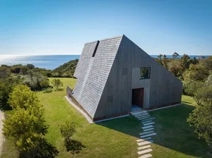 A photo of a triangular house near the ocean with a wooden structure, surrounded by grass and trees
