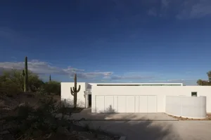 Exterior view of a modern white house with a large garage, two cacti, and a large window, bathed in sunlight