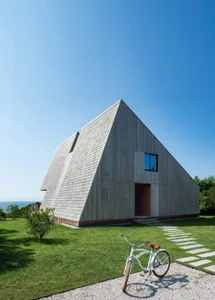 A unique triangular house with a wooden exterior and interior, well-lit with natural sunlight, featuring a bench and two bicycles outside