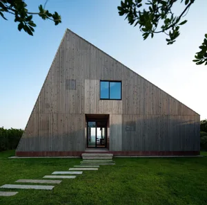 A unique triangular house with a wooden structure, featuring two windows and a chair near one of them, surrounded by a garden area