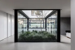 A comfortable indoor space with a glass wall, potted plants, and seating areas, filled with natural light
