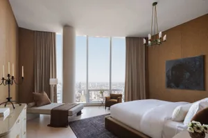 Cozy bedroom with brown walls, a white bed, and a city skyline view through a large window, accompanied by a potted plant and two chairs