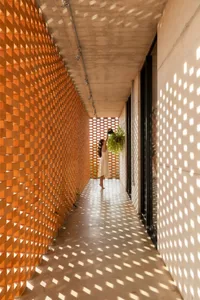 A woman stands in a white tile hallway with a potted plant on the wall, looking at it while holding a pole, with sunlight illuminating the space through windows and two sets of doors visible