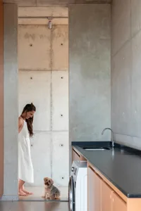 A woman stands in a kitchen with a sink, oven, and refrigerator, surrounded by casual, lived-in decor