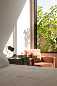 Cozy bedroom with a large window, two chairs, a couch, and a pink and grey chair, with books and a vase adding to the inviting atmosphere