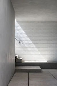 A minimalist staircase with a handrail leads to a white brick wall, set against a possibly residential or commercial building exterior