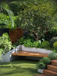 A wooden bench surrounded by greenery and potted plants, with a fence and two bottles in a relaxing outdoor patio setting