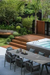 A relaxing outdoor patio area with a wooden deck, hot tub, and dining table surrounded by chairs, surrounded by lush greenery and potted plants