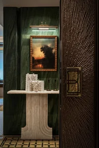 A sophisticated hallway with a marble table, a green wall, a glass door, and a painting above the table, with two candles adding to the ambiance