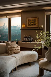 A cozy living room with two couches, a chair, dining table, and decorative items, featuring a large window for natural light and artistic vases, with a warm and inviting atmosphere