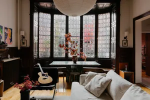 Modern living room with a couch, chairs, dining table, potted plants, and a vase, filled with natural light from large windows