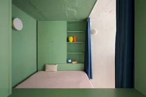 A small bedroom with two beds, a bookshelf, and decorative vases, featuring green walls and a pink bedspread