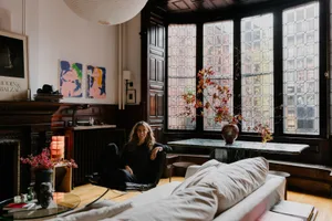 A cozy living area with a woman on a couch in front of a large window, surrounded by a chair, potted plant, and decorative vase, with natural light creating a warm ambiance
