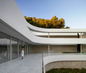 A modern building with a curved design and large circular structure, featuring multiple windows and a woman walking nearby, set in a well-lit outdoor environment
