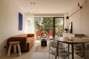 Open living area with couches, chairs, and a dining table, surrounded by greenery and brightly lit