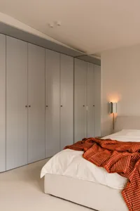 A well-lit bedroom with a large bed, two dressers, and a chair, featuring white walls and a red comforter