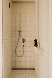 Small bathroom with white tile walls, a white tiled shower stall, and a silver shower head, with a bottle on the right side of the shower area
