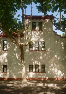 A photo of an old building with red brick walls, white and red elements, and a tree in front of it