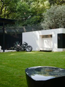 A relaxed outdoor area with a motorcycle parked on a grassy lawn in front of a white building, featuring two benches and a pleasant atmosphere