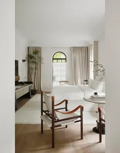 A well-lit bedroom with a white bed, two chairs, a potted plant, and a vase, creating a cozy atmosphere