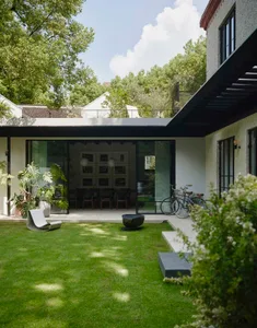 Open living area with a glass door leading to a backyard patio, featuring a dining table, chairs, bicycles, and a kitchen area with a sink and potted plant, bathed in natural light