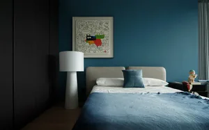 Cozy bedroom with a white bed, blue comforter, wooden floor, and a painting above the bed, lit by a bedside lamp
