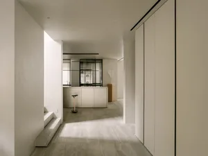 Open living area with white walls and floors, featuring a kitchen, dining, and living space with two chairs, a refrigerator, and a vase