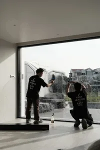 Two men working together to clean windows in a room with potted plants and a collaborative atmosphere