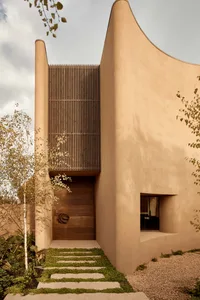 A modern house with curved walls, a wooden door, and two windows on either side, surrounded by a contemporary and stylish living environment