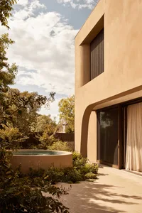 A modern house with a large window in front of a tan wall, surrounded by trees and a pool in a warm climate setting