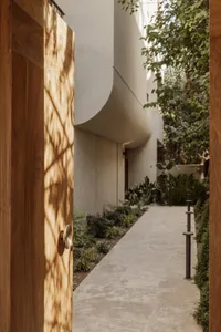 A serene outdoor space with a large white wall, a wooden-framed doorway, and potted plants, accompanied by two benches for seating