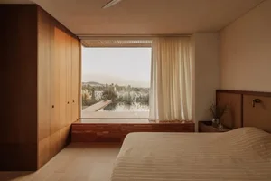 A well-lit bedroom with a bed near a large window, two potted plants, and a vase, creating a cozy and inviting atmosphere