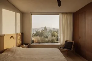 Cozy bedroom with a large window offering a city view, featuring a bed, chair, and two couches, with natural light creating a warm ambiance