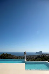 A woman stands on a deck overlooking a large body of water, with a pool nearby, surrounded by a serene atmosphere