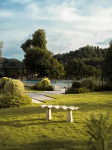 A serene outdoor space with a pool surrounded by grass and trees, featuring two benches and a person sitting on one of them