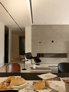 A modern living space with a dining table, chairs, and a couch, surrounded by concrete walls, with natural light and a cozy atmosphere