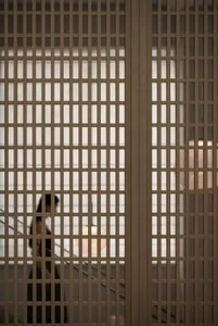 A woman walks down a dimly lit hallway with a white wall and a wooden fence, lit by two lights on either side