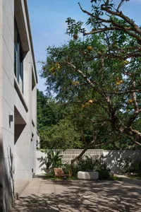A serene outdoor space with a white building, patio, and surrounding trees, featuring a bench and two chairs for relaxation