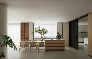 Modern kitchen with wooden cabinets, white walls, and a large window allowing natural light to fill the space, featuring two dining tables and several chairs, with a potted plant adding greenery