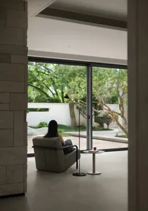 A woman sits in a chair in front of large windows, surrounded by a calm and relaxing atmosphere with a dining table, a potted plant, and two chairs in a bright and inviting space