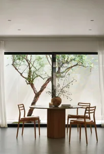 A modern dining space with a wooden table, three chairs, and a vase, surrounded by large windows allowing natural sunlight to enter