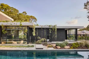 A modern house with a black concrete exterior, surrounded by trees and a pool, featuring potted plants and two chairs for relaxation