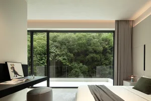 Modern bedroom with white walls, a black bed, and a large window overlooking a forest, with a calm and serene ambiance