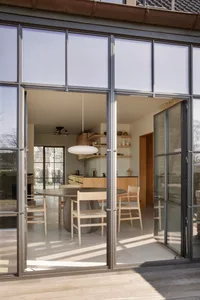 Open modern kitchen and dining space with wooden cabinets, a large round dining table, and comfortable seating arrangement, surrounded by natural light