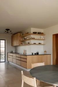 Open kitchen with wooden cabinets, white walls, a sink, dining table, chairs, and various bowls and cups
